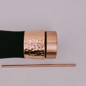 May include: A black water bottle with a hammered copper band and cap, and a copper straw. The bottle has a sleek design, with a textured band adding visual interest. The straw is thin and straight.