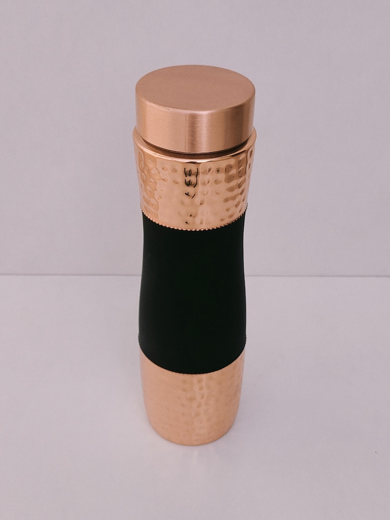 May include: A copper-coloured water bottle with a hammered texture on the top and bottom sections. The middle section is black. The bottle has a matching copper lid. The bottle is cylindrical and appears to be made of metal.