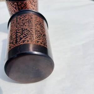 May include: Two copper-coloured metal tumblers with intricate floral patterns. The rims and outer base are black. The interior of the tumblers is copper. The tumblers are cylindrical and appear to be handmade.