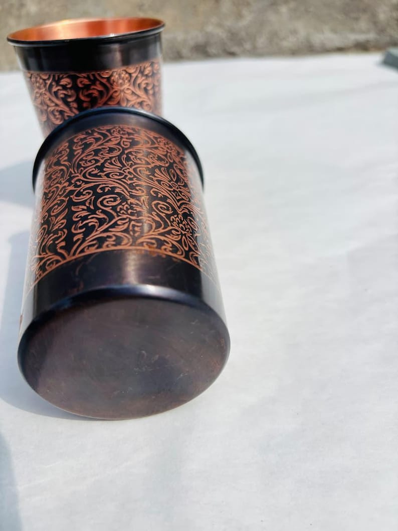 May include: Two copper-coloured metal tumblers with intricate floral patterns. The rims and outer base are black. The interior of the tumblers is copper. The tumblers are cylindrical and appear to be handmade.