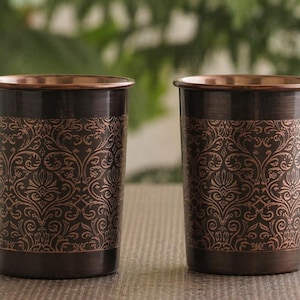 May include: Two copper-coloured drinking glasses with a dark brown base and rim. The glasses feature an ornate, repeating floral pattern in a lighter copper hue. The background is blurred, with green foliage visible.