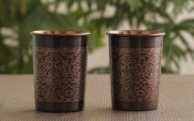 May include: Two copper-coloured drinking glasses with a dark brown base and rim. The glasses feature an ornate, repeating floral pattern in a lighter copper hue. The background is blurred, with green foliage visible.