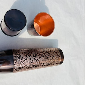 May include: Three copper-coloured metal tumblers with a decorative floral pattern. One tumbler is upside down, revealing a dark brown base. The other two tumblers are upright, showcasing the copper interior. The design is a mix of dark brown and copper.