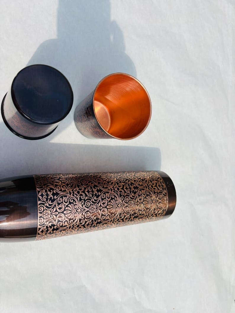 May include: Three copper-coloured metal tumblers with a decorative floral pattern. One tumbler is upside down, revealing a dark brown base. The other two tumblers are upright, showcasing the copper interior. The design is a mix of dark brown and copper.