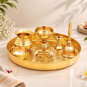 May include: A polished gold-tone metal puja thali set featuring a round tray with an embossed swastika symbol, a small water pot, a bowl, a diya lamp, a small stand, and a tall hand bell. The items are arranged together on the circular tray.