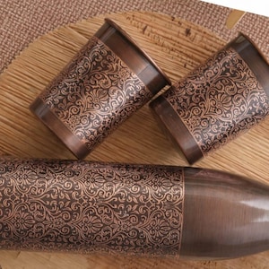 May include: A copper-coloured water bottle and two matching cups with an ornate floral pattern. The items are arranged on a wooden surface, with a woven mat in the background. The bottle has a screw-on cap.