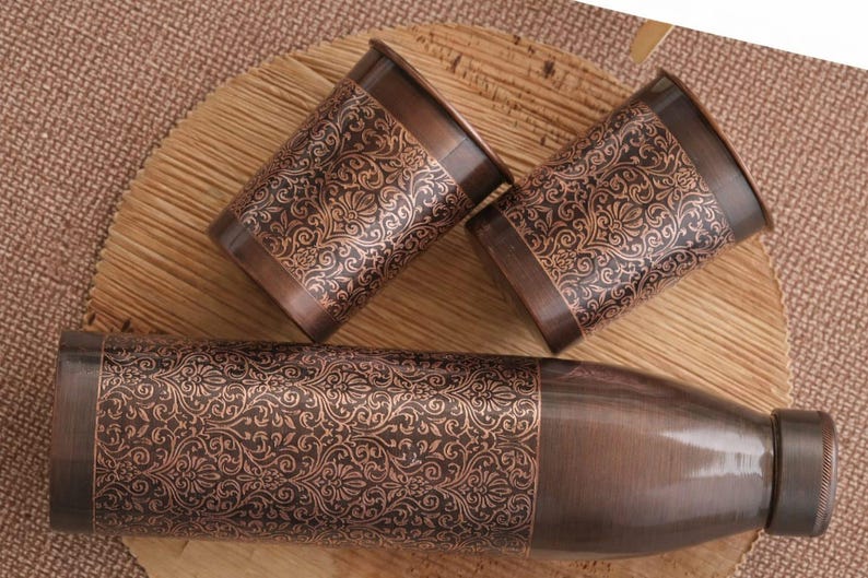 May include: A copper-coloured water bottle and two matching cups with an ornate floral pattern. The items are arranged on a wooden surface, with a woven mat in the background. The bottle has a screw-on cap.