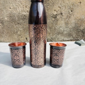 May include: A copper-toned water bottle and two matching tumblers. The bottle and tumblers feature a dark brown base with an ornate, repeating floral pattern in a lighter copper hue. The interior of the tumblers is a bright copper colour.