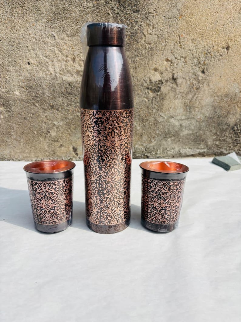 May include: A copper-toned water bottle and two matching tumblers. The bottle and tumblers feature a dark brown base with an ornate, repeating floral pattern in a lighter copper hue. The interior of the tumblers is a bright copper colour.