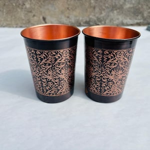 May include: Two copper-coloured drinking glasses. The exterior is black with an ornate, swirling floral pattern, while the interior is bright copper. The cylindrical glasses have a slightly flared rim. Each glass is approximately 10 cm tall.