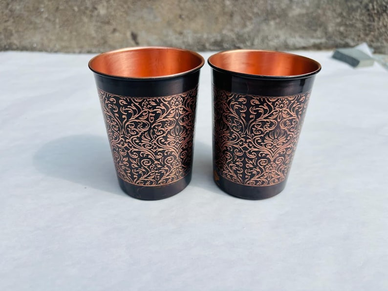 May include: Two copper-coloured drinking glasses. The exterior is black with an ornate, swirling floral pattern, while the interior is bright copper. The cylindrical glasses have a slightly flared rim. Each glass is approximately 10 cm tall.