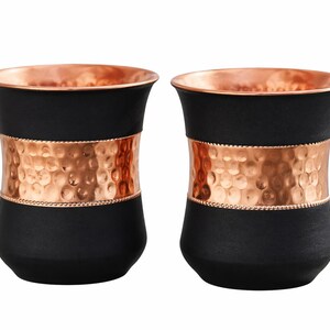 May include: Two black drinking glasses with copper-colored rims and a hammered copper band around the middle. The glasses have a sleek, modern design, perfect for serving beverages.