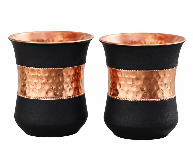 May include: Two black drinking glasses with copper-colored rims and a hammered copper band around the middle. The glasses have a sleek, modern design, perfect for serving beverages.