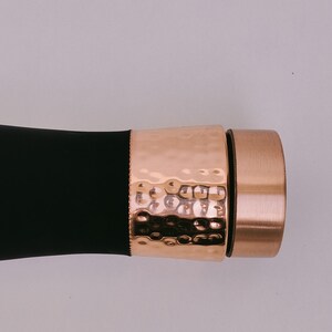 May include: A black object with a hammered copper-colored band and a copper-colored cap. The band has a textured, dimpled surface. The cap is a solid copper color. The object is set against a light background.