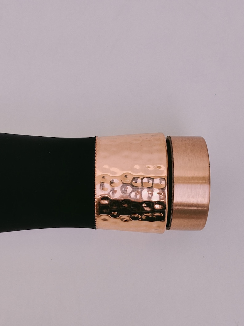 May include: A black object with a hammered copper-colored band and a copper-colored cap. The band has a textured, dimpled surface. The cap is a solid copper color. The object is set against a light background.