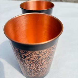 May include: Two copper-coloured metal cups with a black exterior and an ornate, swirling pattern. The interior of the cups is a bright copper colour. The cups are likely for drinking and have a simple, elegant design.