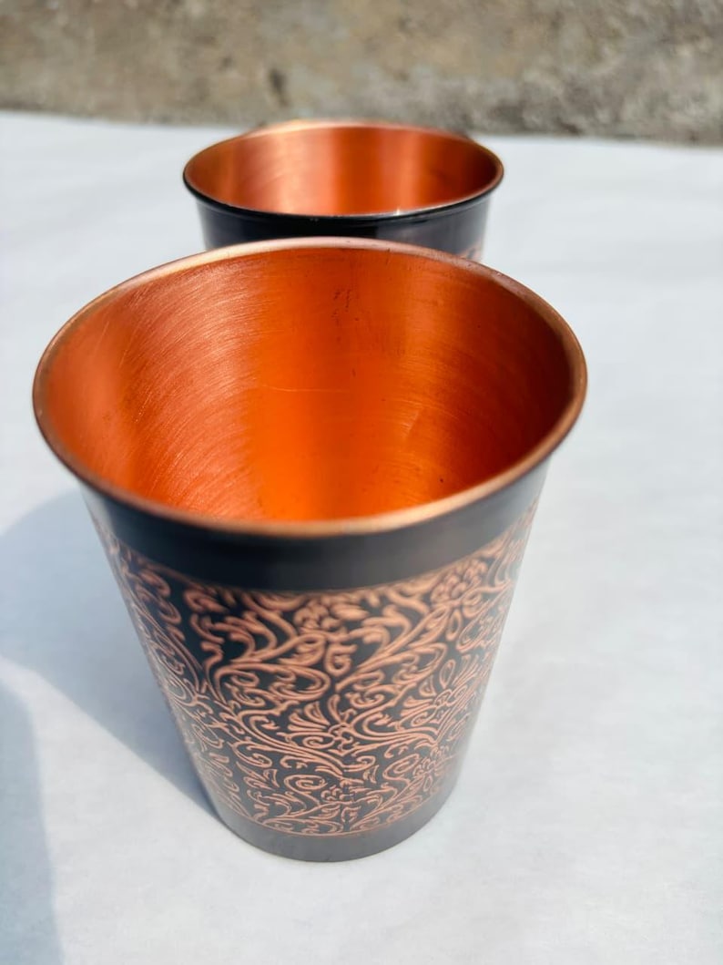 May include: Two copper-coloured metal cups with a black exterior and an ornate, swirling pattern. The interior of the cups is a bright copper colour. The cups are likely for drinking and have a simple, elegant design.