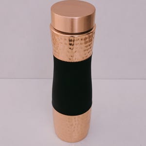 May include: A copper-colored water bottle with a hammered texture on the top and bottom sections. The middle section is black. The bottle has a matching copper lid. The design is sleek and modern.
