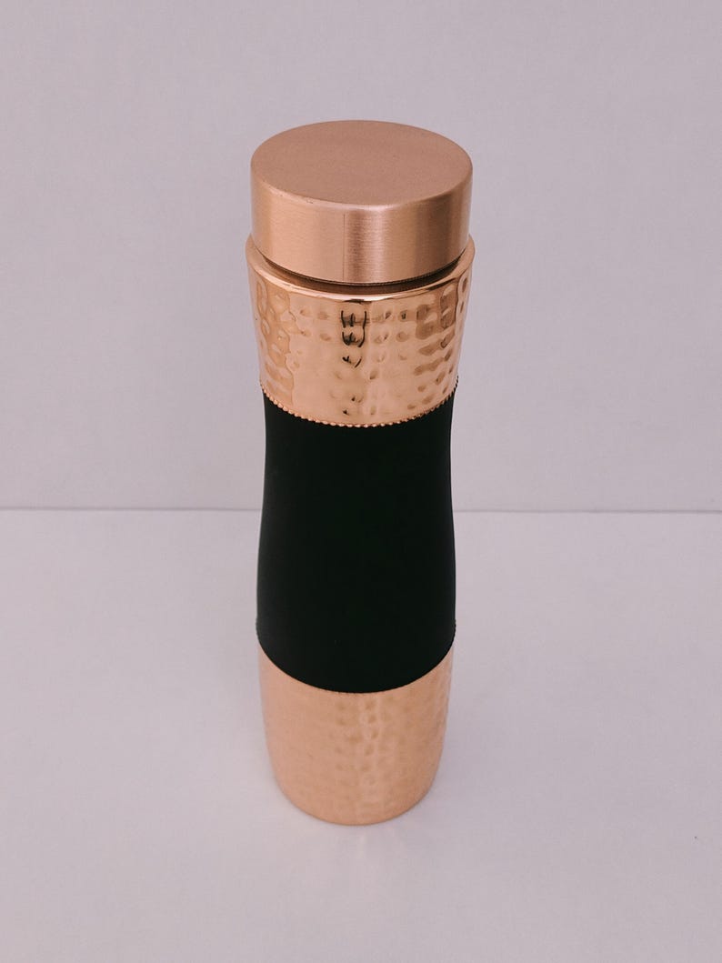 May include: A copper-colored water bottle with a hammered texture on the top and bottom sections. The middle section is black. The bottle has a matching copper lid. The design is sleek and modern.