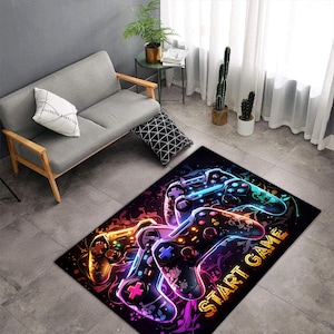 Puede incluir: Una alfombra rectangular que presenta un gr&aacute;fico vibrante de colores ne&oacute;n de varios mandos de videojuegos sobre un fondo oscuro y abstracto. El dise&ntilde;o incluye acentos brillantes en naranja, azul y morado con el texto START GAME en amarillo.