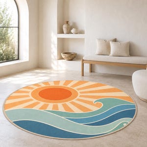 May include: A circular area rug featuring a graphic design of a bright orange sun with radiating light rays positioned above stylized ocean waves in shades of light blue, medium blue, and dark blue, all set against a cream-colored background.
