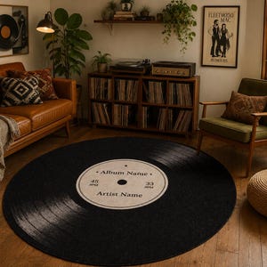 May include: A circular area rug designed to look like a black vinyl record. The center label is beige with black text that reads Album Name, Artist Name, 45 RPM, and 33 RPM. The rug features concentric grooves and a central hole graphic.