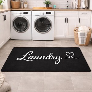 May include: Black rectangular laundry room rug with the word "Laundry" in white script and a heart symbol. The rug is placed on a tiled floor in front of a washing machine and dryer.