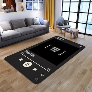 May include: A black rectangular rug designed to look like a music player interface. The rug has a large black screen with the text "ALBUM COVER HERE" and playback controls. The rug is placed on a wooden floor in a living room setting.