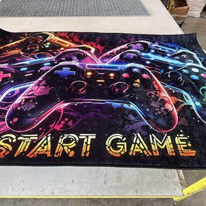 Puede incluir: Una alfombra rectangular con fondo negro y un estampado gr&aacute;fico multicolor de tres mandos de videojuegos. El dise&ntilde;o incluye remolinos de ne&oacute;n en rosa, azul y naranja, con el texto START GAME impreso en color amarillo.