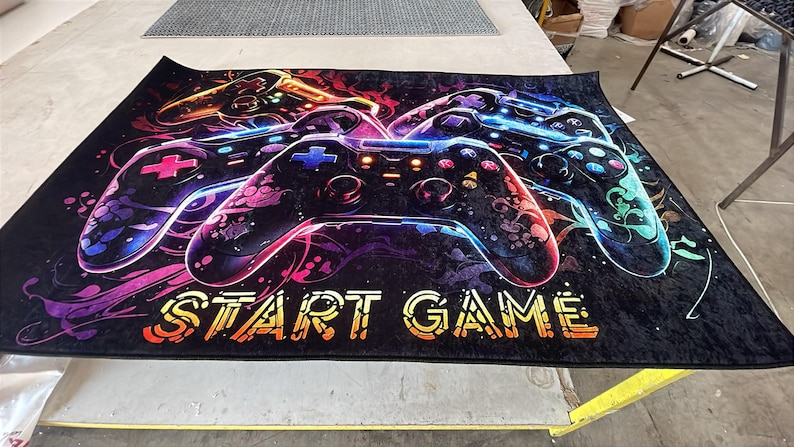 Puede incluir: Una alfombra rectangular con fondo negro y un estampado gr&aacute;fico multicolor de tres mandos de videojuegos. El dise&ntilde;o incluye remolinos de ne&oacute;n en rosa, azul y naranja, con el texto START GAME impreso en color amarillo.