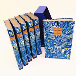 May include: A set of books with blue and white marbled covers. The spines have brown labels with text. A blue box is behind the books. The book on the right has the title "The Mathnawi I" and the author's name.