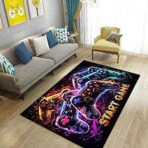 Puede incluir: Una alfombra rectangular con fondo negro y un estampado gr&aacute;fico vibrante de varios mandos de videojuegos en colores ne&oacute;n. El dise&ntilde;o incluye patrones abstractos en espiral y el texto START GAME en letras de color amarillo.
