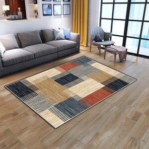 May include: A rectangular area rug with a geometric pattern. The rug features blocks of color in beige, navy blue, rust, and gray, with a black border. The design includes both solid and striped sections.