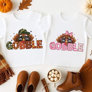 May include: Two white toddler t-shirts with turkey graphics and the word "GOBBLE" in plaid letters. One turkey wears a camo hat and sunglasses, the other a pink bow. The image includes brown shoes, a scarf, and pumpkins.