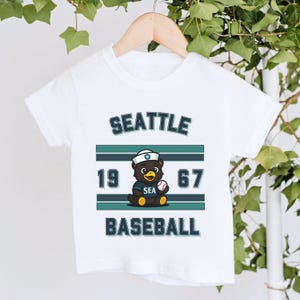 May include: A white short-sleeve t-shirt featuring a graphic of a cartoon bear wearing a sailor hat and holding a baseball. The text reads SEATTLE, 1967, SEA, and BASEBALL, accompanied by horizontal teal and dark blue stripes on the front.