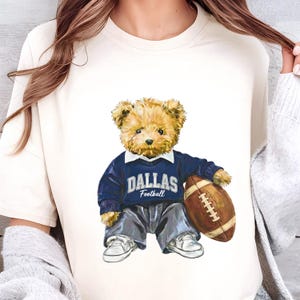 May include: Cream-colored t-shirt featuring a teddy bear wearing a navy blue sweater with "DALLAS Football" text. The bear holds a brown football and is dressed in gray pants and white sneakers.