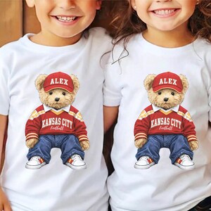 Puede incluir: Dos camisetas blancas con un dise&ntilde;o de oso de peluche. El oso lleva una gorra roja con "ALEX", un su&eacute;ter rojo y amarillo con "KANSAS CITY Football", vaqueros azules y zapatillas blancas con detalles rojos y amarillos.