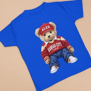 Puede incluir: Camiseta azul real con un oso de peluche que lleva una gorra roja con "ALEX", un su&eacute;ter rojo y blanco de Kansas City Football, vaqueros azules y zapatillas blancas. El oso es el foco principal.