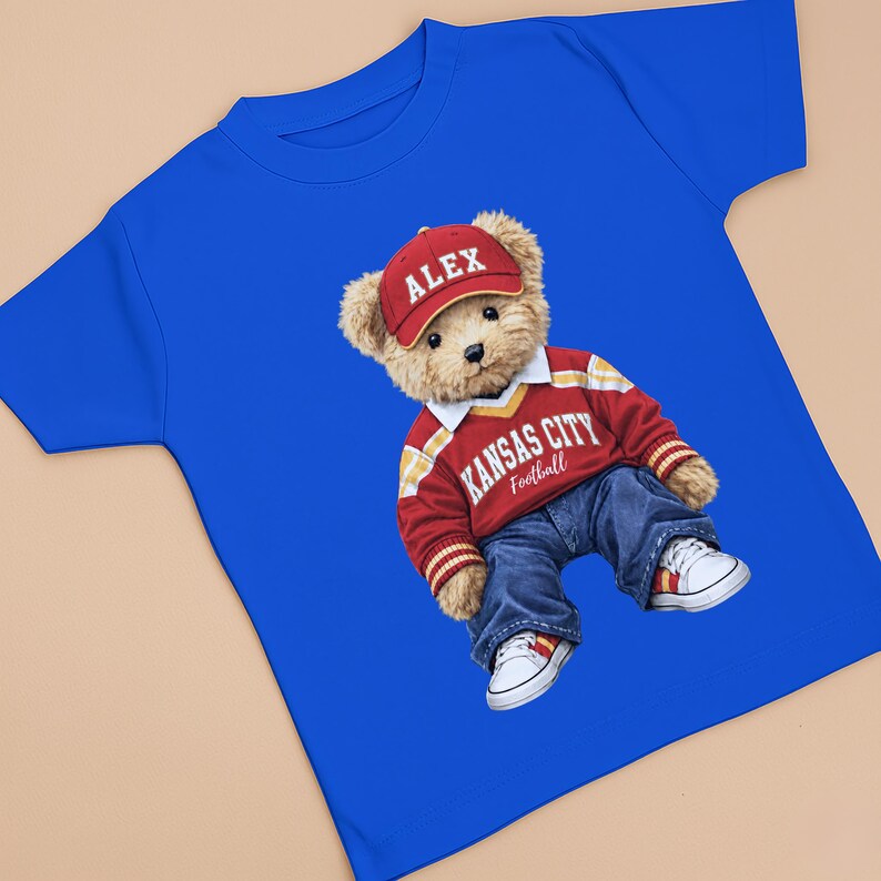 Puede incluir: Camiseta azul real con un oso de peluche que lleva una gorra roja con "ALEX", un su&eacute;ter rojo y blanco de Kansas City Football, vaqueros azules y zapatillas blancas. El oso es el foco principal.