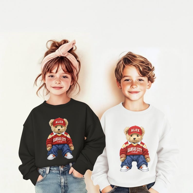 Puede incluir: Dos ni&ntilde;os con sudaderas. Uno lleva una negra, el otro una blanca. Ambos tienen un dibujo de un oso de peluche con gorra roja y chaqueta roja y blanca con "Kansas City Football". El oso tambi&eacute;n lleva vaqueros azules y zapatillas blancas.