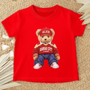 Puede incluir: Camiseta roja con un oso de peluche que lleva una gorra roja con "ALEX", un su&eacute;ter rojo y blanco con "KANSAS CITY Football", vaqueros azules y zapatillas blancas. El gr&aacute;fico del oso de peluche est&aacute; centrado en la camiseta.