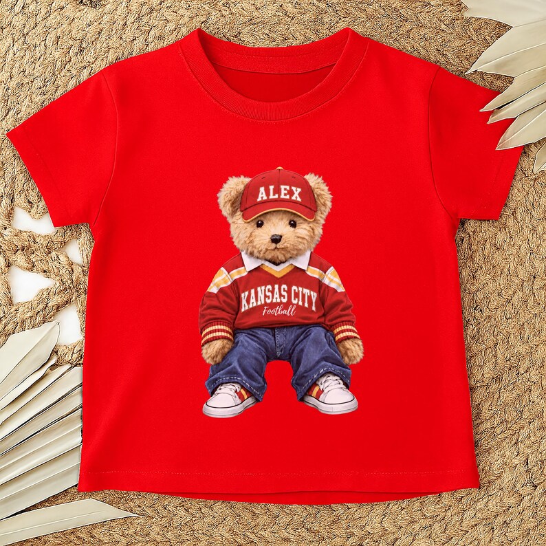 Puede incluir: Camiseta roja con un oso de peluche que lleva una gorra roja con "ALEX", un su&eacute;ter rojo y blanco con "KANSAS CITY Football", vaqueros azules y zapatillas blancas. El gr&aacute;fico del oso de peluche est&aacute; centrado en la camiseta.