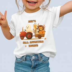 Puede incluir: Camiseta blanca con un gráfico colorido de un tractor rojo tirando de un vagón lleno de animales de dibujos animados y calabazas. El texto "AMY'S FALL ADVENTURE BEGINS!" está impreso en naranja. La camiseta la lleva un niño.