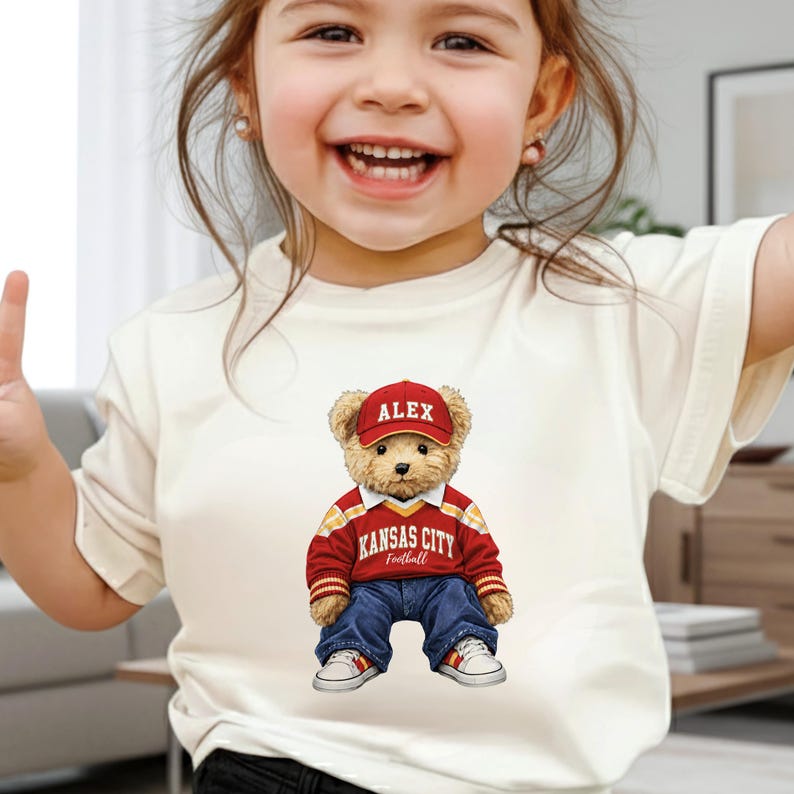 Puede incluir: Camiseta color crema con un gr&aacute;fico de un oso de peluche que lleva una gorra roja con "ALEX", una camiseta de f&uacute;tbol roja y dorada de Kansas City, vaqueros azules y zapatillas blancas. El oso est&aacute; centrado en la camiseta.