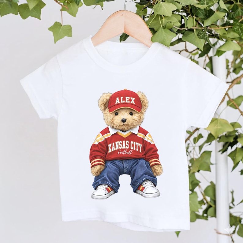 Puede incluir: Camiseta blanca con un oso de peluche que lleva una gorra roja con "ALEX", un su&eacute;ter rojo y amarillo con "KANSAS CITY Football", vaqueros azules y zapatillas blancas. La camiseta est&aacute; colgada en una percha de madera.