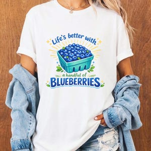 May include: White t-shirt with a colorful graphic of a carton of blueberries. The text reads "Life's better with a handful of BLUEBERRIES" in blue and yellow. The shirt is worn by a person.