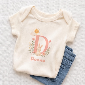 May include: Cream-colored baby onesie with a pink letter "D" decorated with a bunny, flowers, and a sun. The name "Donna" is printed below the letter. Blue denim jeans are partially visible.