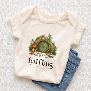 Könnte beinhalten: Cremefarbener Baby-Body mit einer skurrilen Illustration einer grünen Hobbit-Tür in einem grasbewachsenen Hügel, mit Pilzen und dem Wort "halfling" darunter. Blaue Jeans sind unter dem Body gefaltet.