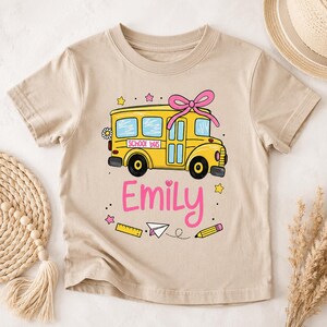 Op de afbeelding: Een beige t-shirt met korte mouwen en een afbeelding van een gele schoolbus met een roze strik op het dak. Op de zijkant van de bus staat de tekst "SCHOOL bus". Onder de bus staat de naam "Emily" in het roze, omringd door een liniaal, potlood en sterren.
