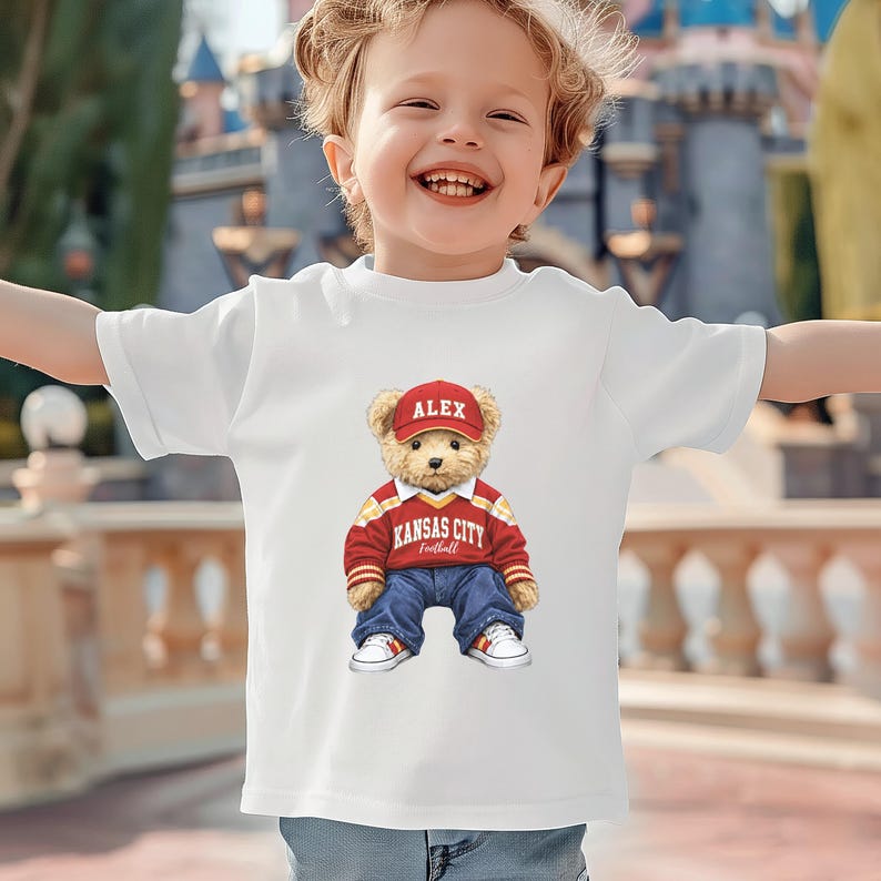 Puede incluir: Camiseta blanca con un oso de peluche de dibujos animados que lleva una gorra roja con "ALEX", un jersey rojo y amarillo con "KANSAS CITY Football", vaqueros azules y zapatillas blancas. La camiseta est&aacute; en un ni&ntilde;o.